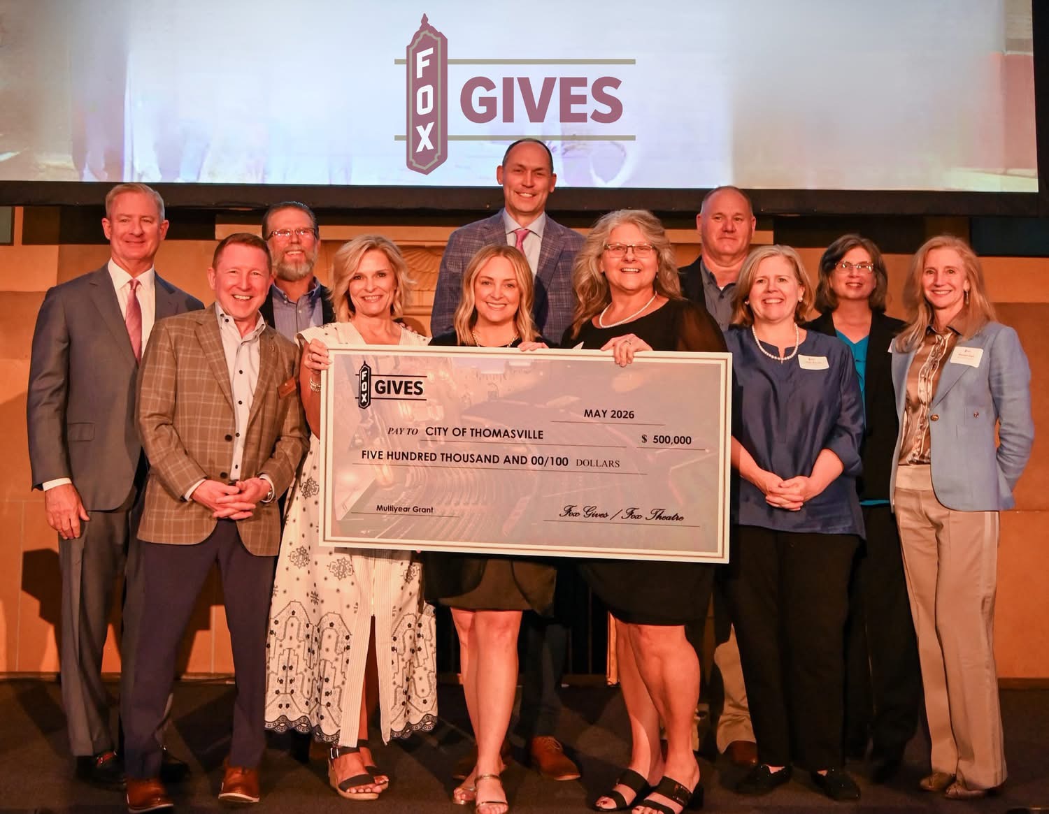 Group of professionals stand on stage holding large check for $500,000 addressed to the City of Thomasville. Fox Gives logo is displayed on a projector behind the group.